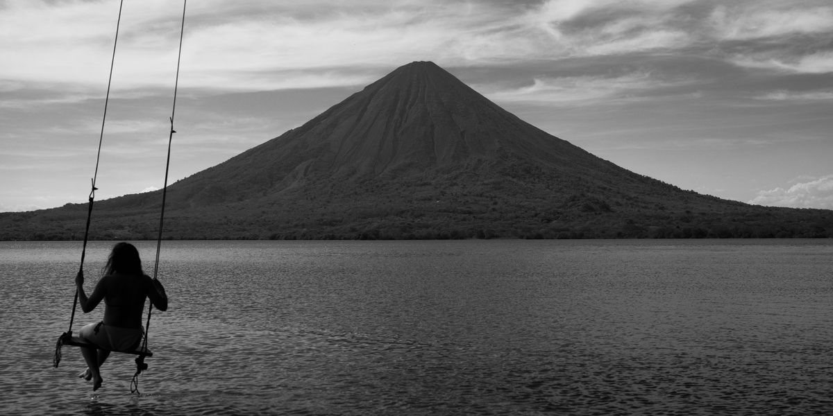 Black-and-white photograph of a person on a swing above still water, a volcano behind them